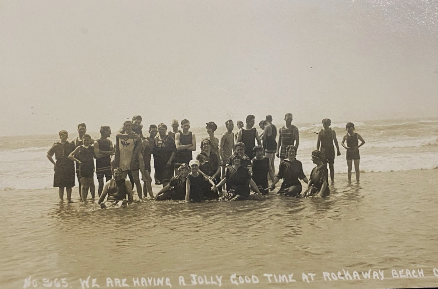Group of people posing in shallow water on a beach, with a handwritten caption along the bottom reading