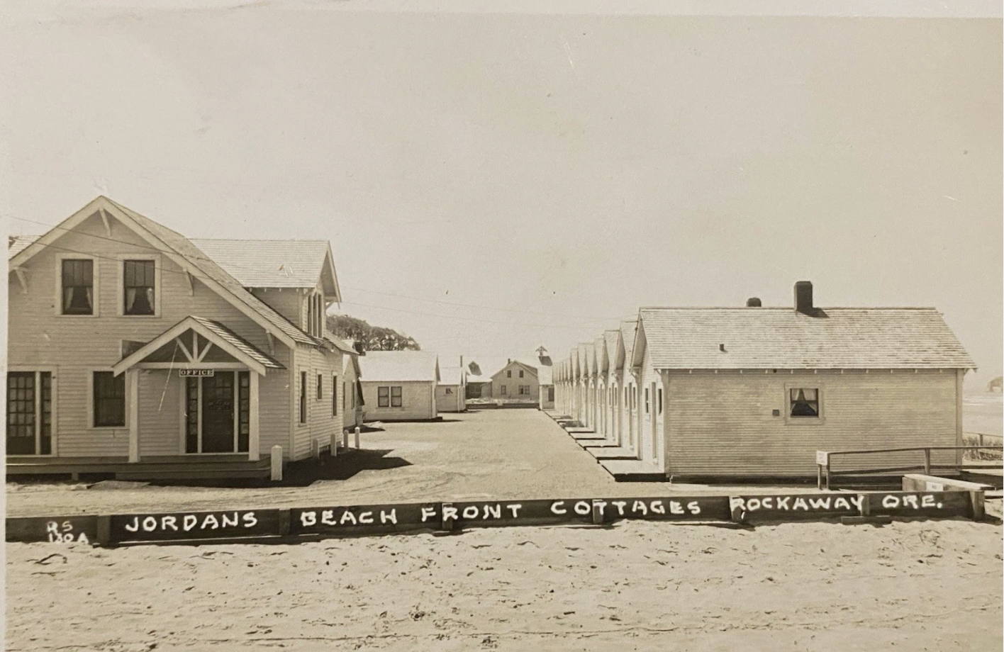 Sepia beach-front cottages lining a sandy path, with a larger house on the left and small sheds on the right.