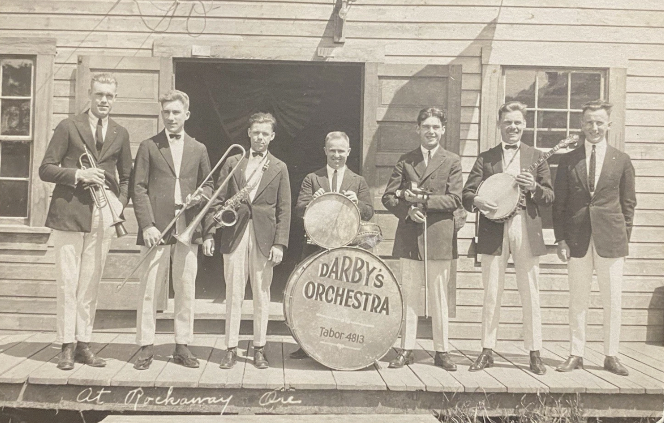 Seven musicians pose on a porch with instruments and a drum labeled “Darby’s Orchestra.”