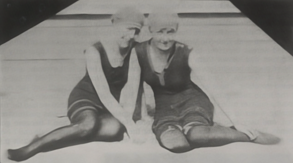 Two seated dancers in 1920s dark swimsuits on a boardwalk, one with a bent knee and one with legs extended.