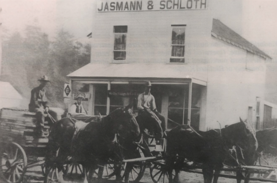 Horse-drawn wagon in front of a Jasmann & Schloth storefront, with people and stacked goods.