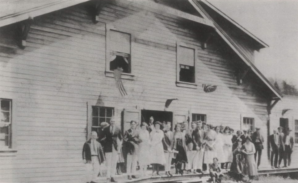 Crowd gathered outside a wooden building, with people standing on the porch and windows above.