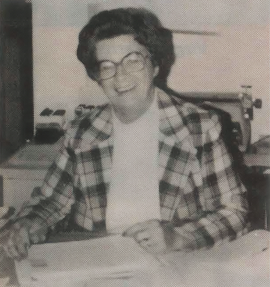 Woman in plaid jacket and glasses seated at a desk, smiling and writing on paper.