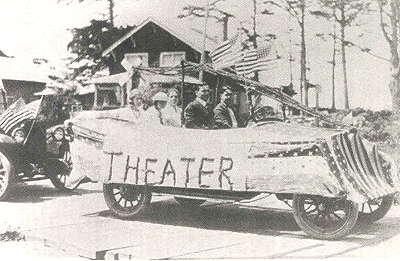 Vintage car with a theater sign on the side, decorated in american flags parked outdoors near a house and trees.