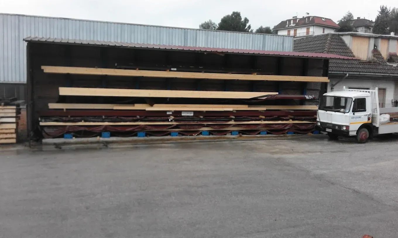 Magazzino industriale con legname accatastato sotto una tettoia e un camion bianco parcheggiato sulla destra.
