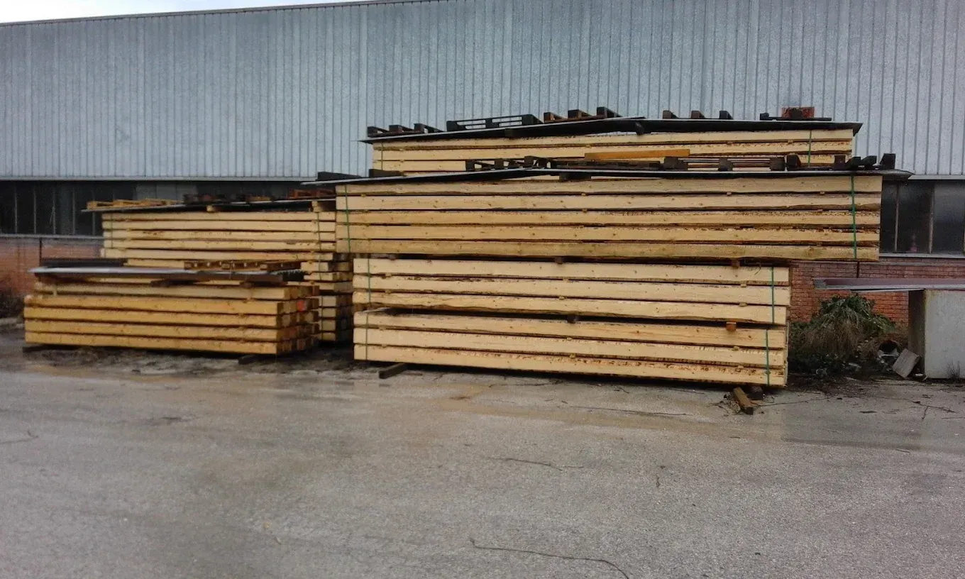 Cataste di assi di legno conservate all’aperto accanto a una parete di lamiera ondulata.