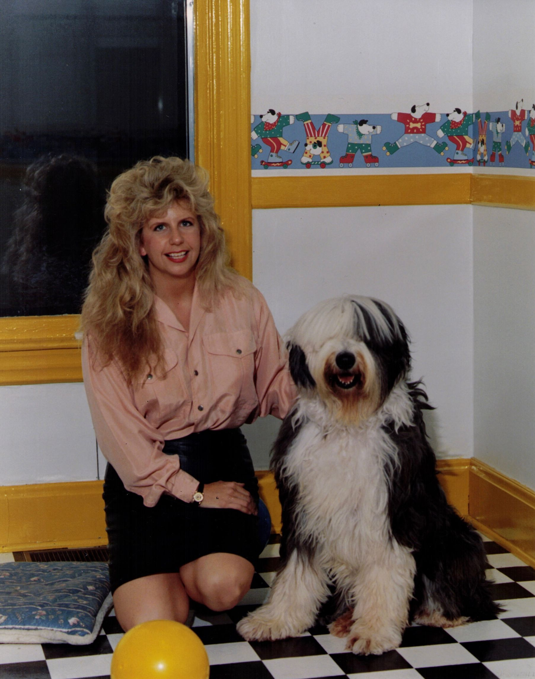 Woman kneeling beside a fluffy black-and-white dog in a colorful room with checkered floor and yellow trim