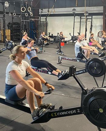 People rowing on Concept2 machines in Great Woods CrossFit, a gym in Norton, MA, with several athletes exercising indoors.