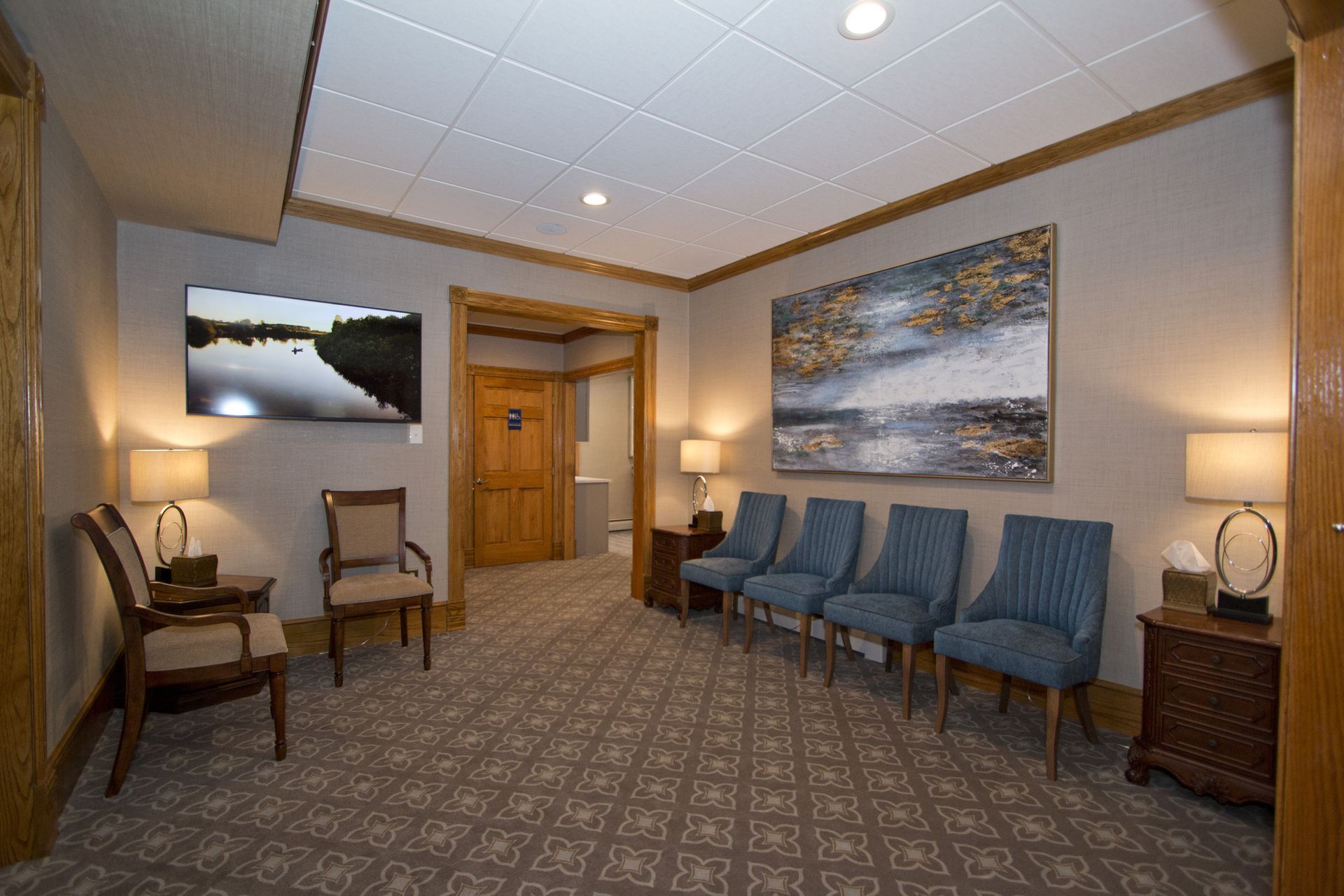 Quiet lobby waiting area with blue chairs, lamps, and wall art in a carpeted room.