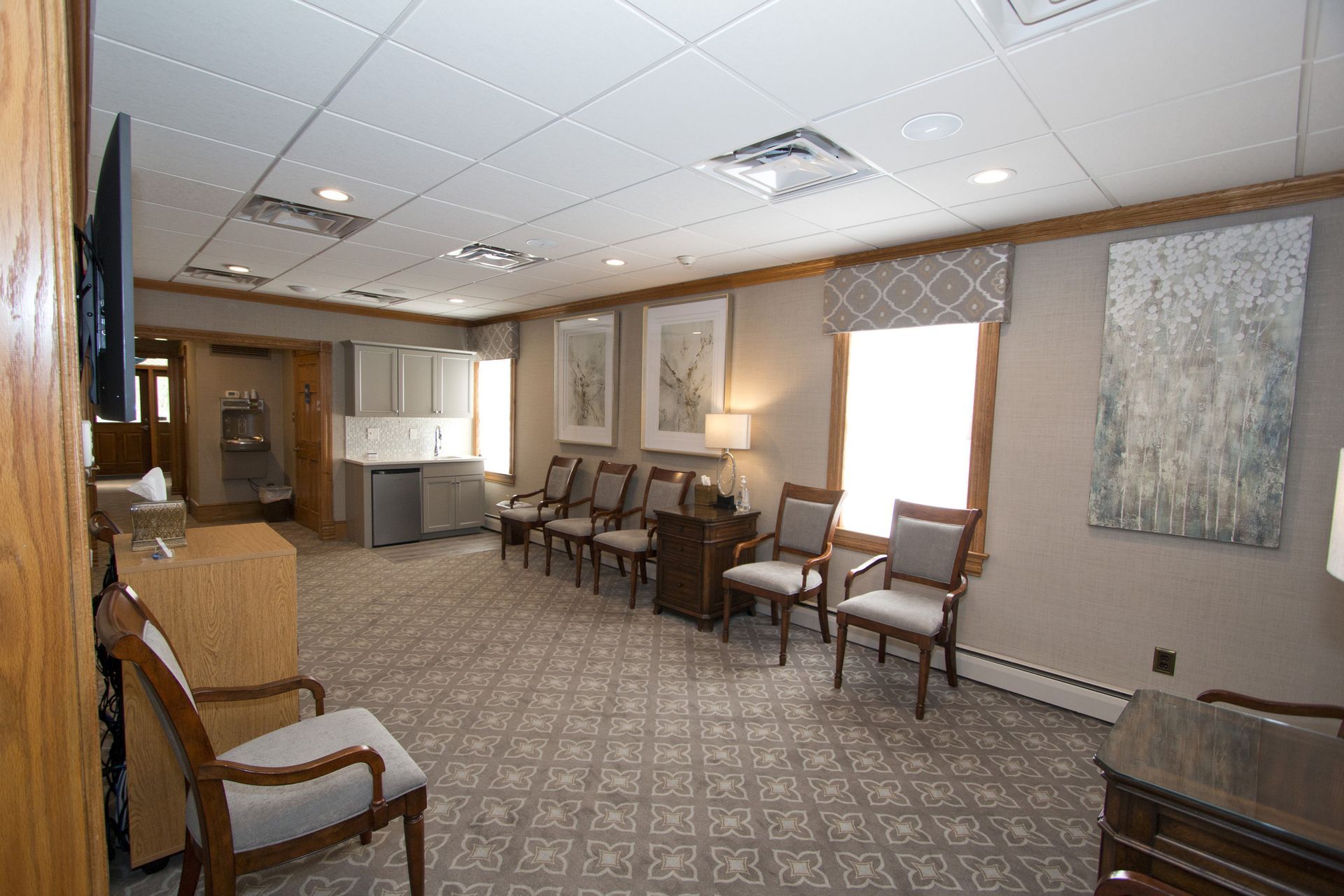 Empty waiting room with chairs, patterned carpet, bright windows, and a wall-mounted TV.