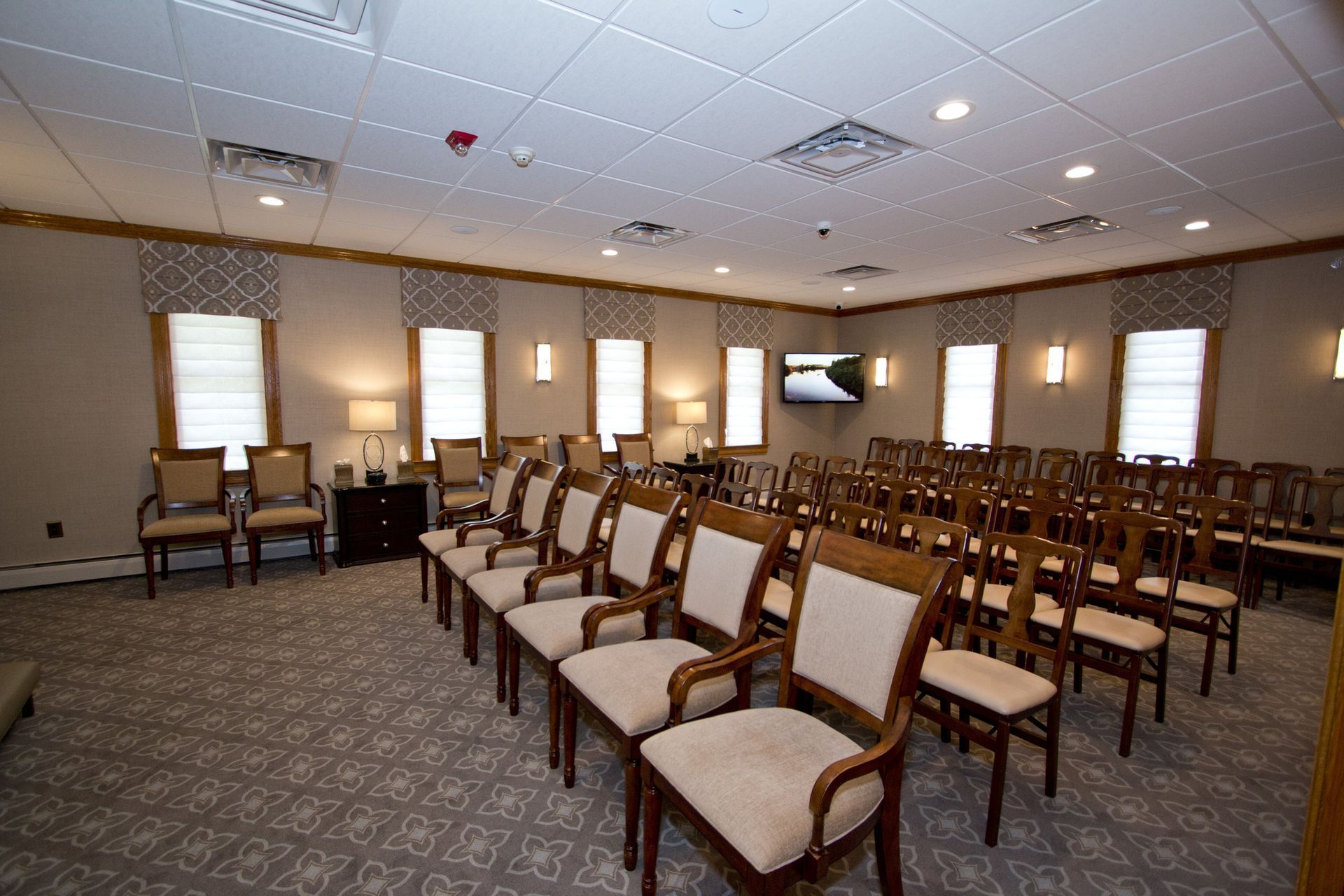 Empty conference room with rows of chairs, patterned carpet, and wall-mounted TV under bright ceiling lights