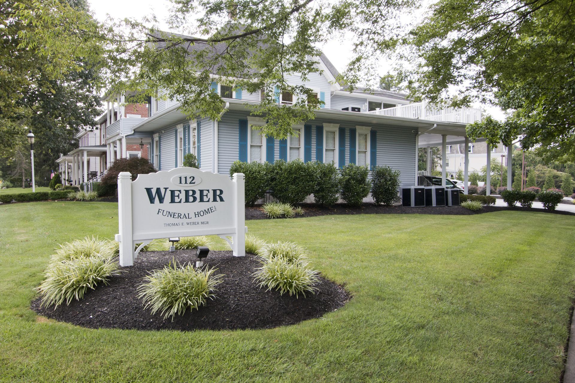 Weber sign in front of a white house with blue shutters and a manicured lawn under trees