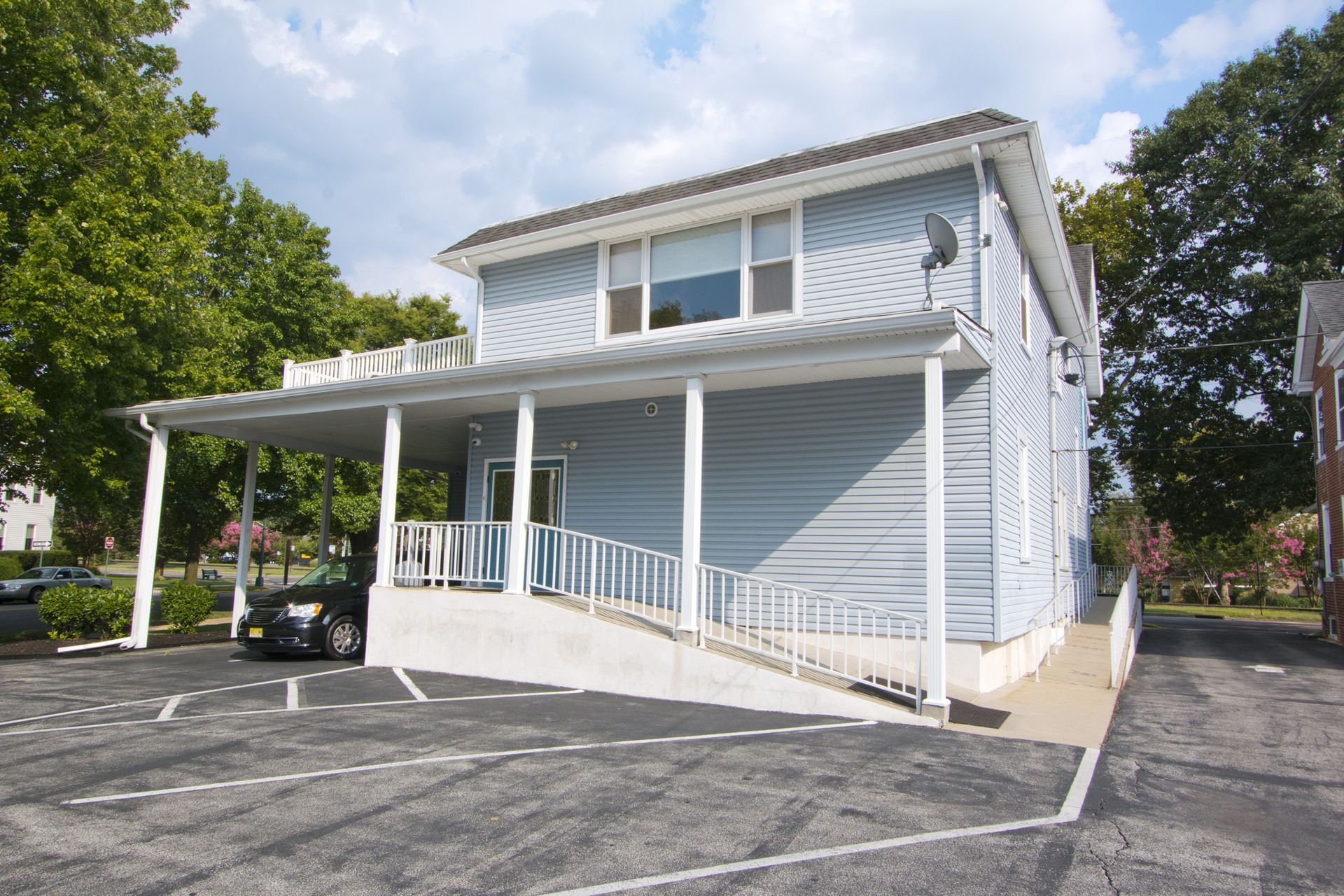 Two-story light blue house with a ramp and covered porch beside a parking lot
