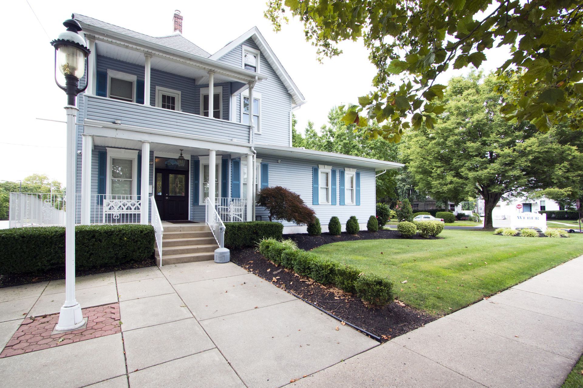 Blue two-story house with front porch, white trim, and a landscaped lawn along a sidewalk