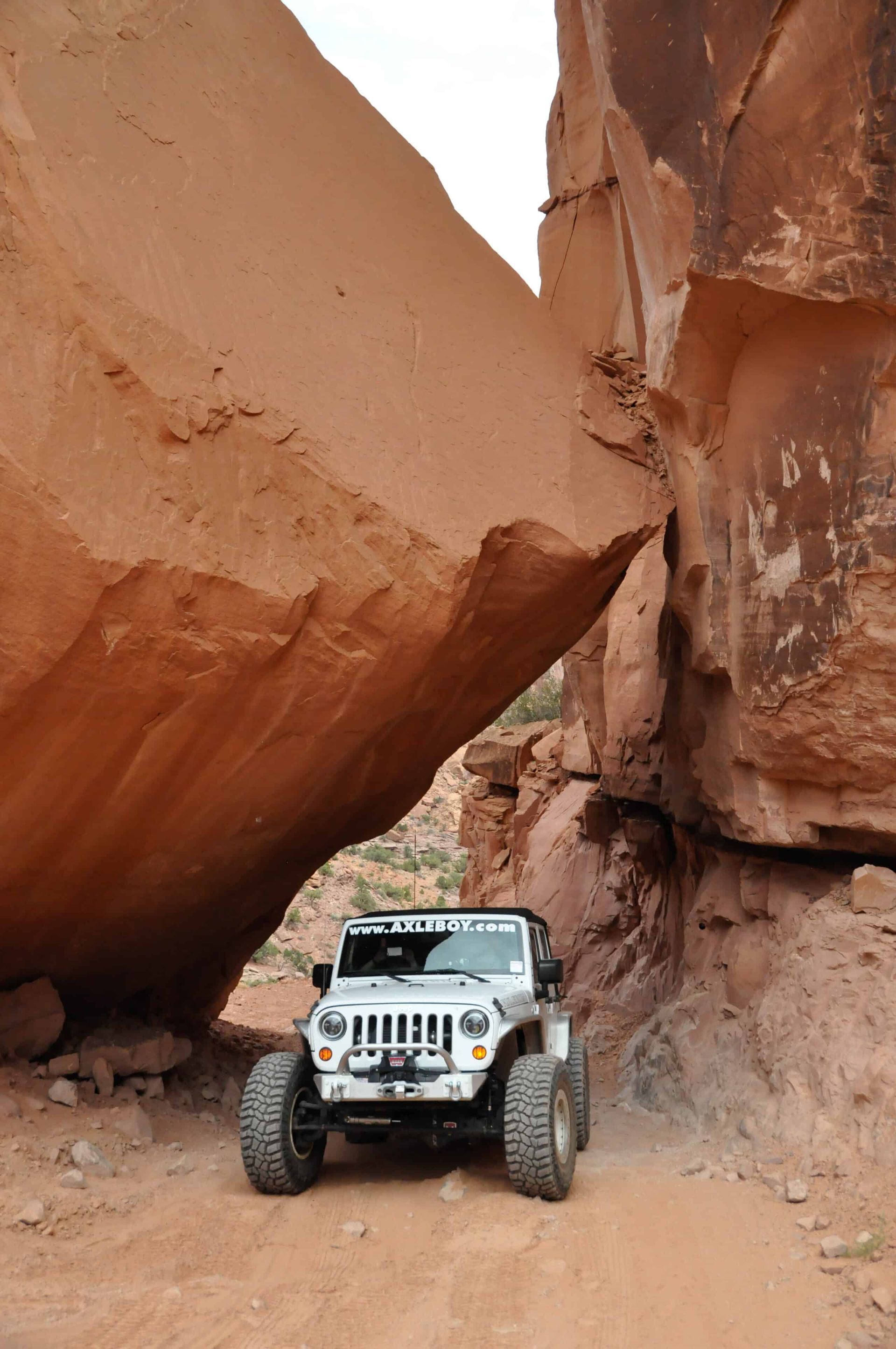 White Jeep off-roading through a narrow canyon, beneath a large reddish-brown rock formation.