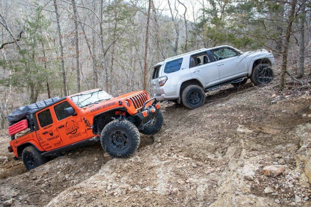 Orange Jeep and silver SUV off-roading on a rocky trail in a wooded area.