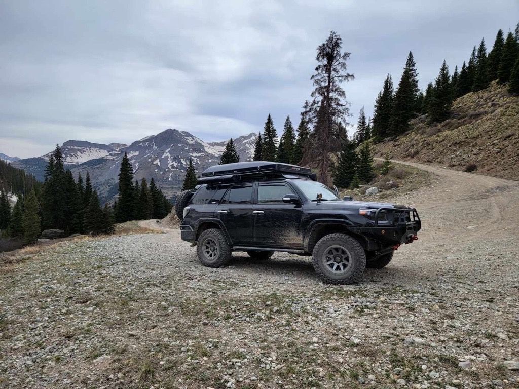 Black SUV on a mountain trail, surrounded by trees and a snowy mountain backdrop.