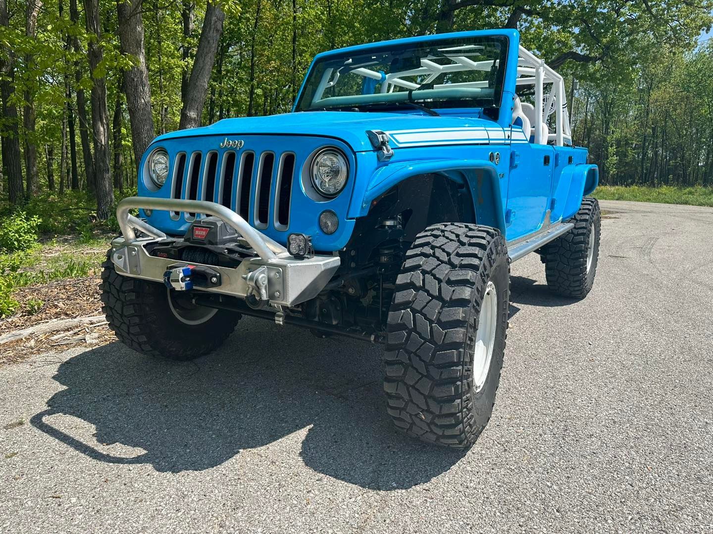 Blue Jeep Wrangler off-road vehicle with white roll cage, rugged tires, and silver bumper parked on pavement.