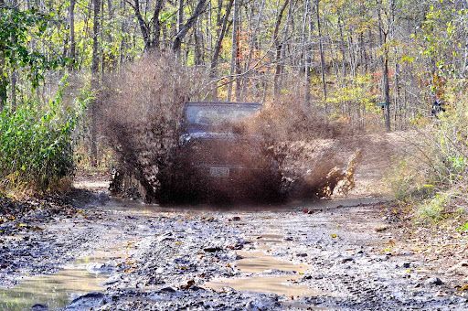 Jeep splashing through muddy water on a forest path, kicking up mud.