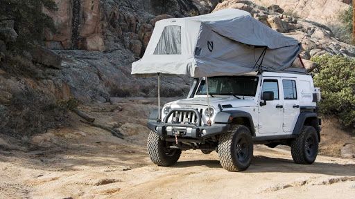 White Jeep Wrangler with a rooftop tent parked on a dirt road in a rocky landscape.