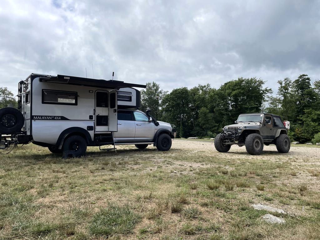 Truck camper and modified Jeep parked outdoors. Grey sky, green trees.