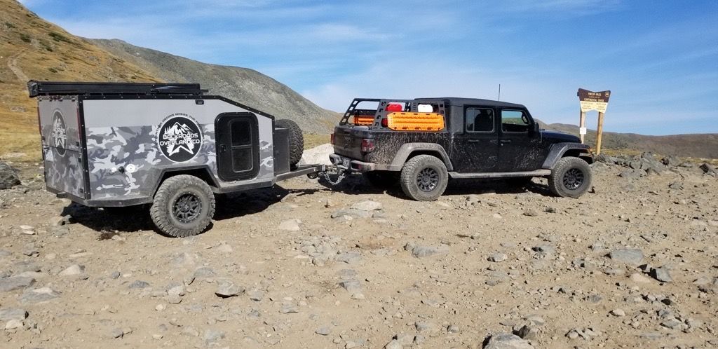 A black Jeep towing a camping trailer on a rocky dirt road in a mountain setting.