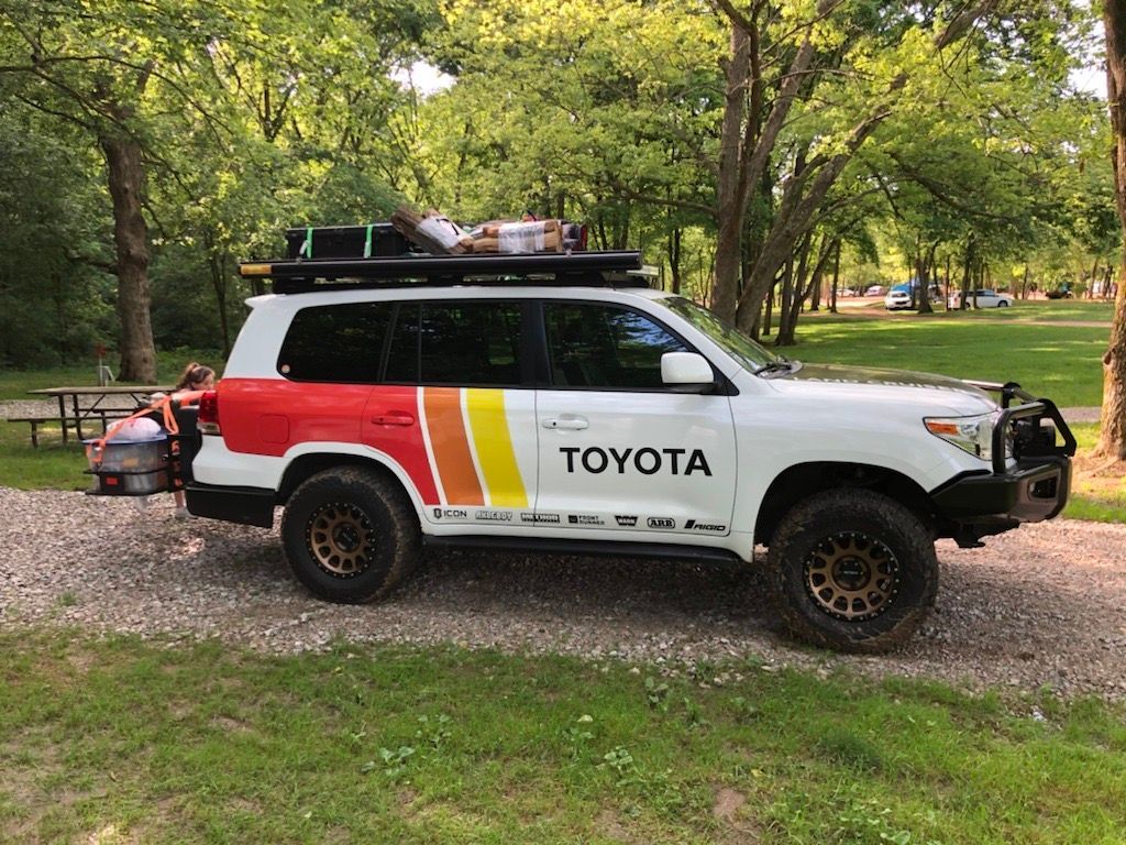 White Toyota SUV with custom red, orange, yellow stripes, roof rack, and off-road tires parked in a grassy campsite.