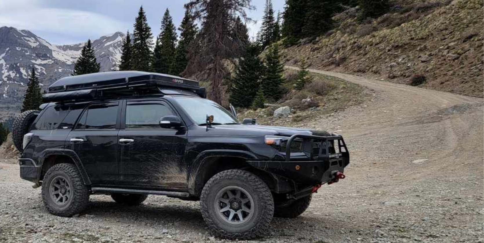 Black SUV on a dirt road in a mountainous area. It has a roof rack, spare tire, and a front bumper.