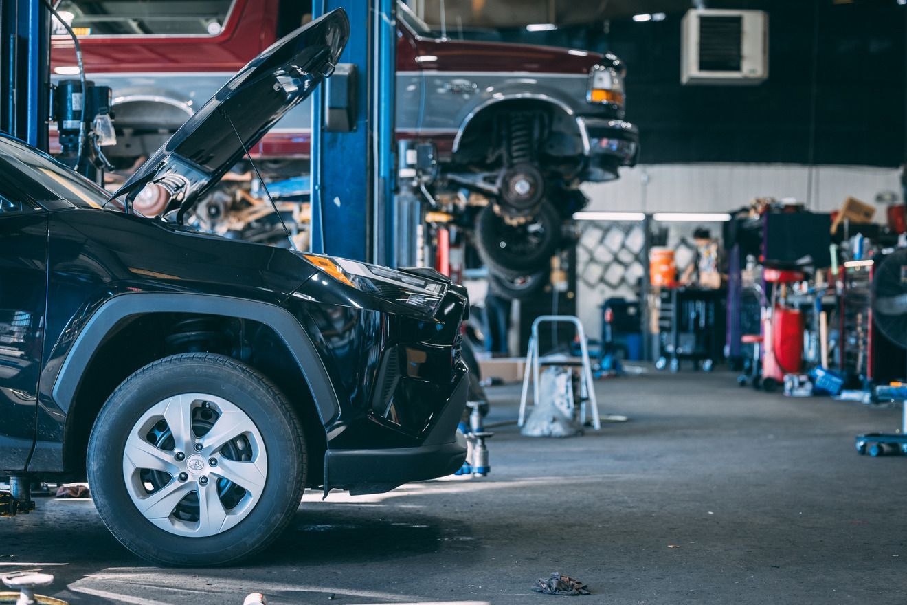 Black SUV in a mechanic shop with the hood open, another car lifted in the background.