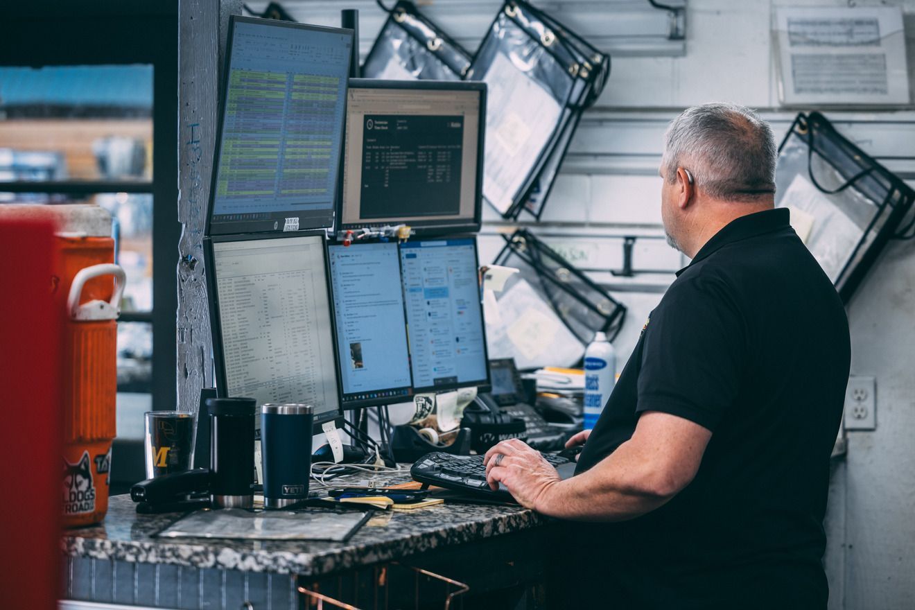 Man working at a cluttered desk with multiple computer screens.  He is looking at the screens, in an indoor setting.