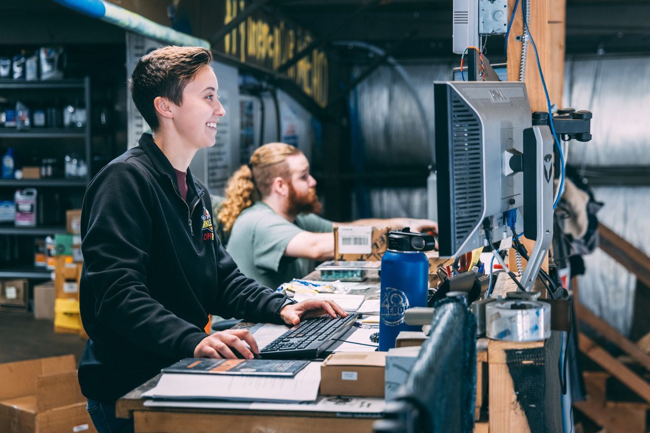 Woman smiles while working at a computer; another person works in the background at a desk in a warehouse.
