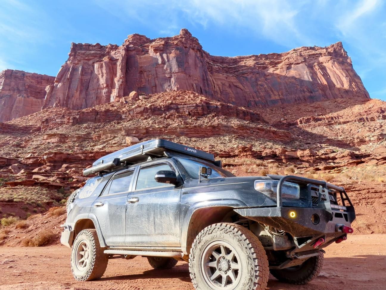 Black SUV with off-road modifications parked in front of a red rock cliff under a blue sky.