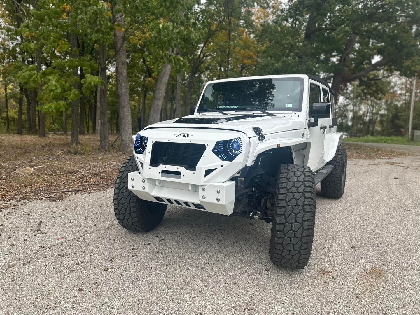 White modified Jeep Wrangler on a gravel road, large tires, custom grill, blue headlights, outdoor setting.