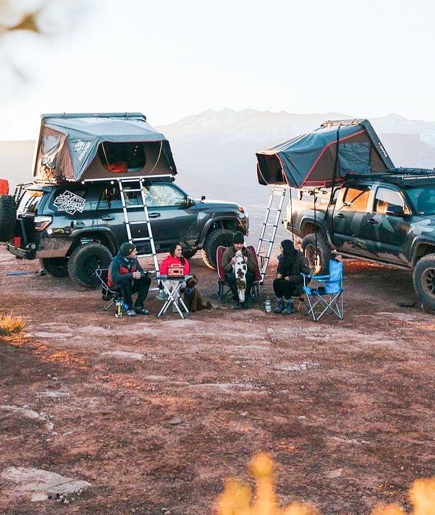 People camping outdoors near two trucks with rooftop tents. Sunset lighting.