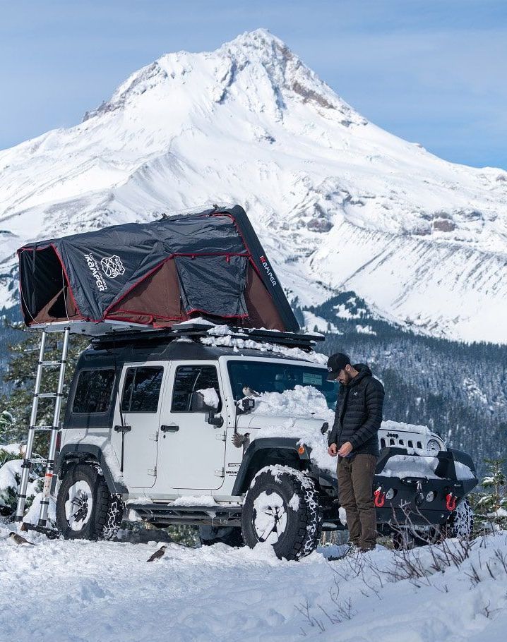 White Jeep with rooftop tent in snow, man standing by. Snowy mountain background.