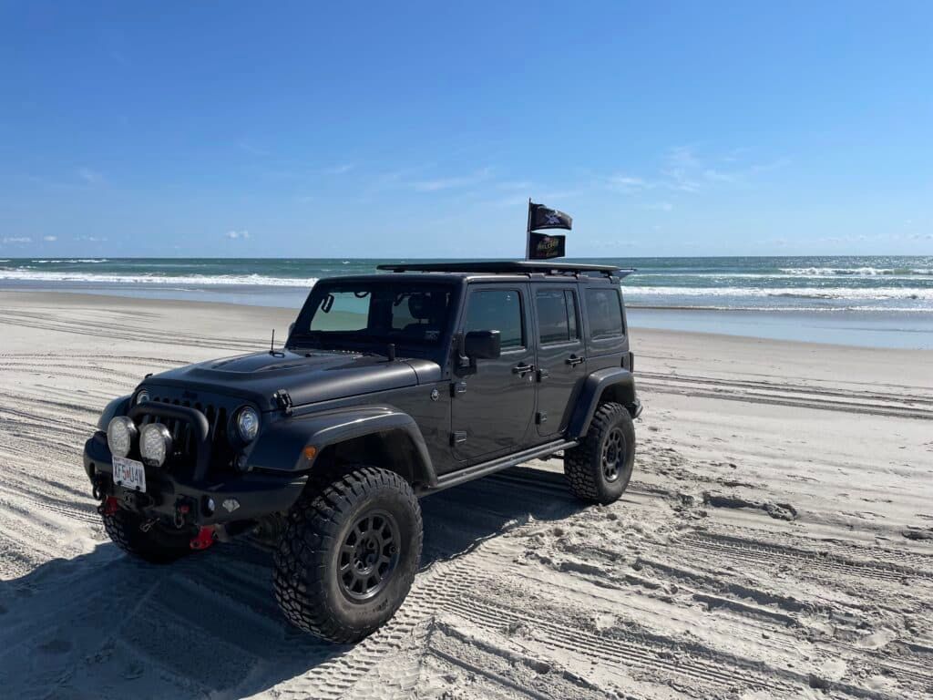 Black Jeep on a sandy beach with the ocean in the background under a blue sky.