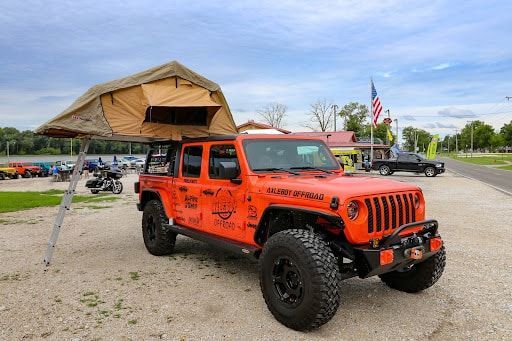 Orange Jeep with rooftop tent parked on gravel. American flag in the background.