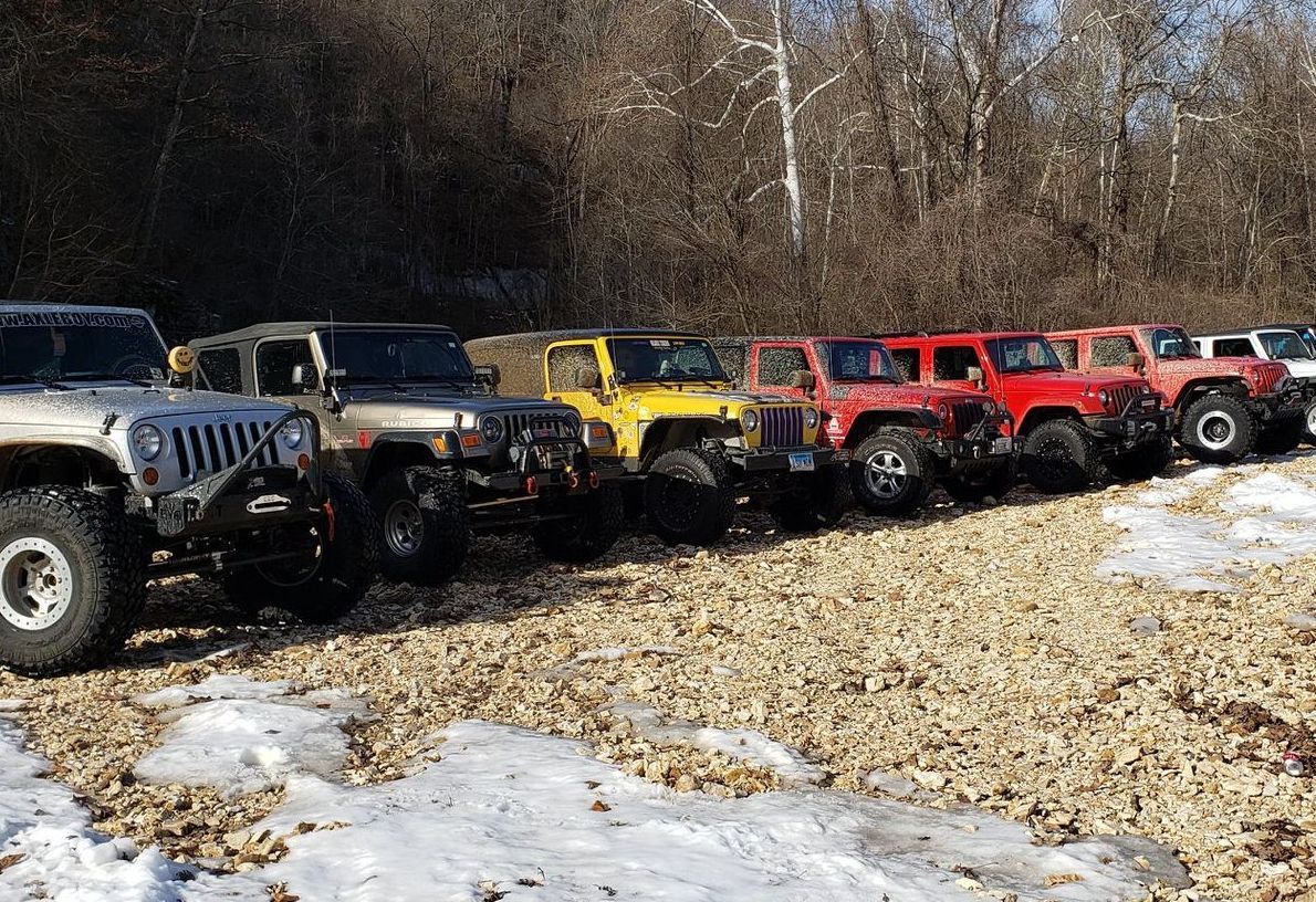 Row of Jeeps of various colors parked on a rocky, snowy terrain near trees.