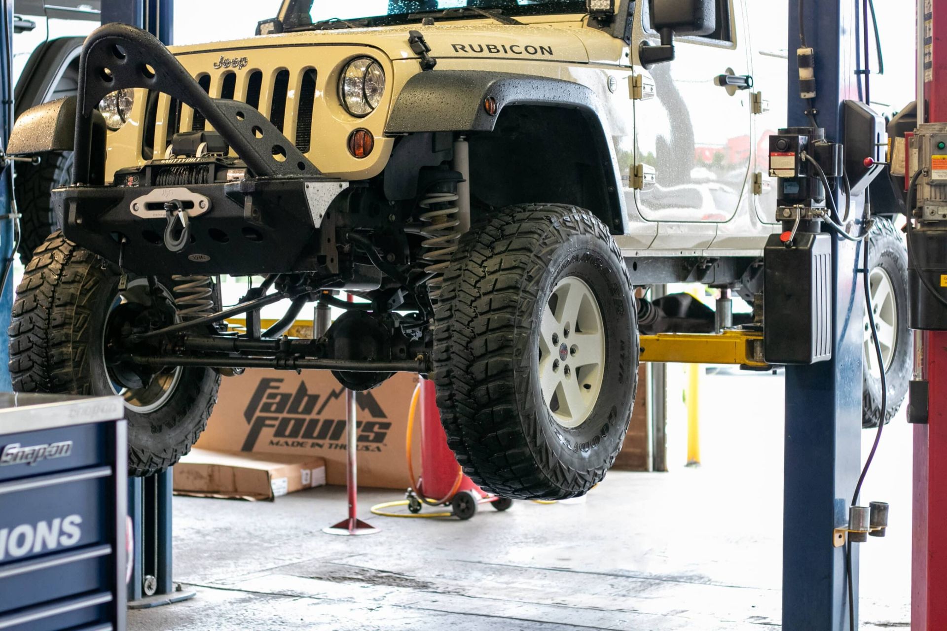 Tan Jeep Rubicon raised on a hydraulic lift in a professional garage.