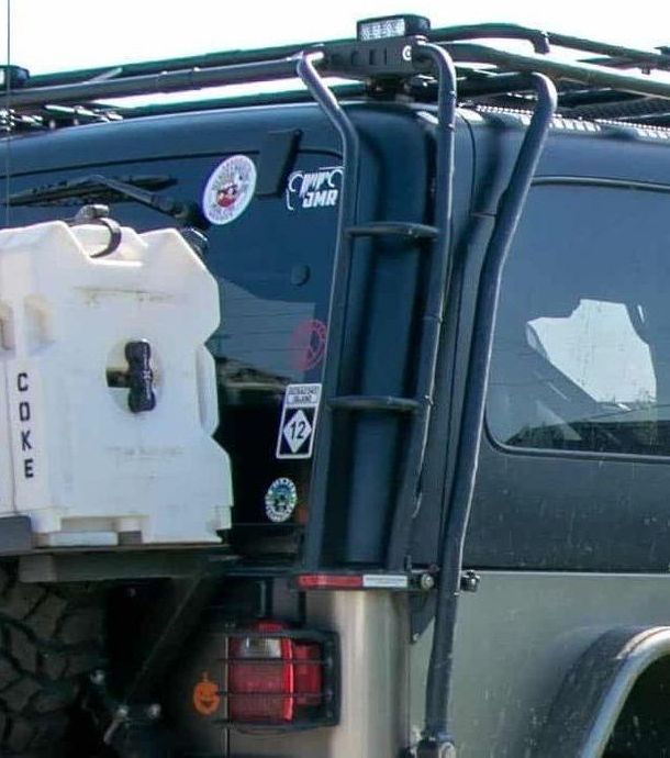Black Jeep with ladder, roof rack, light bar, and water container.