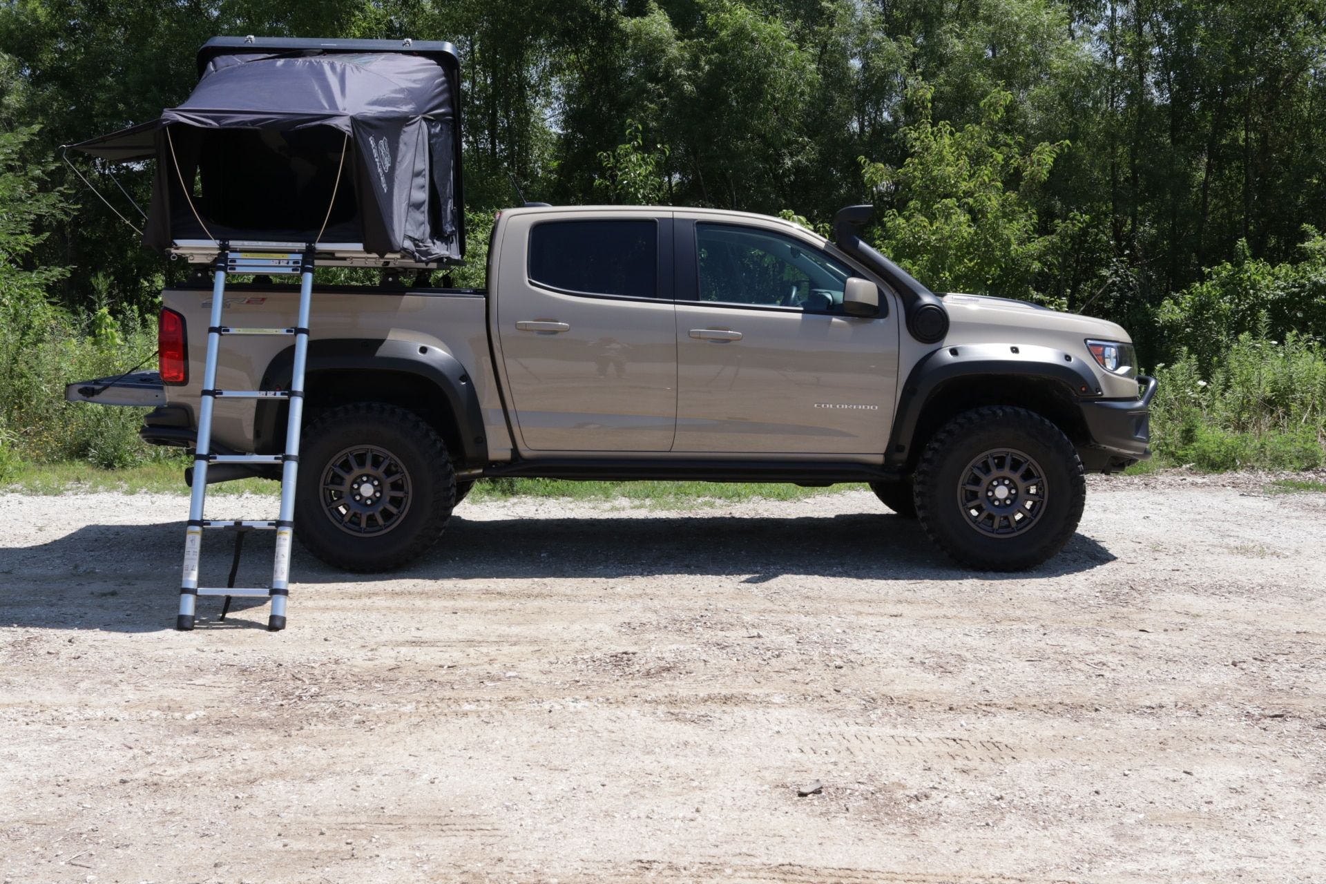 A tan pickup truck with a dark rooftop tent mounted on the bed and a blue aluminum ladder attached