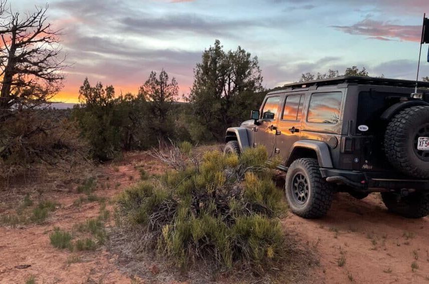 Black Jeep parked on a dirt road at sunset, surrounded by bushes and trees under a colorful sky.