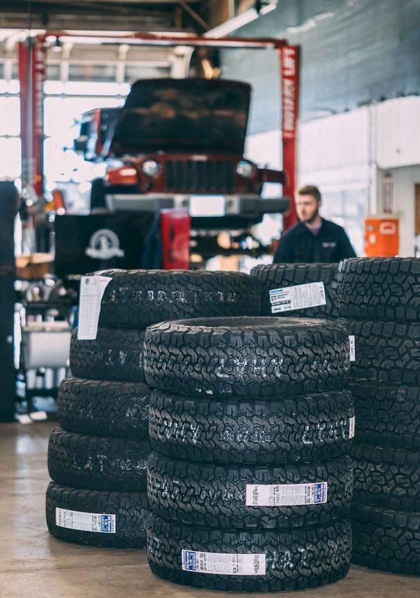 Piles of new tires stacked in a garage, with a car on a lift and a mechanic in the background.