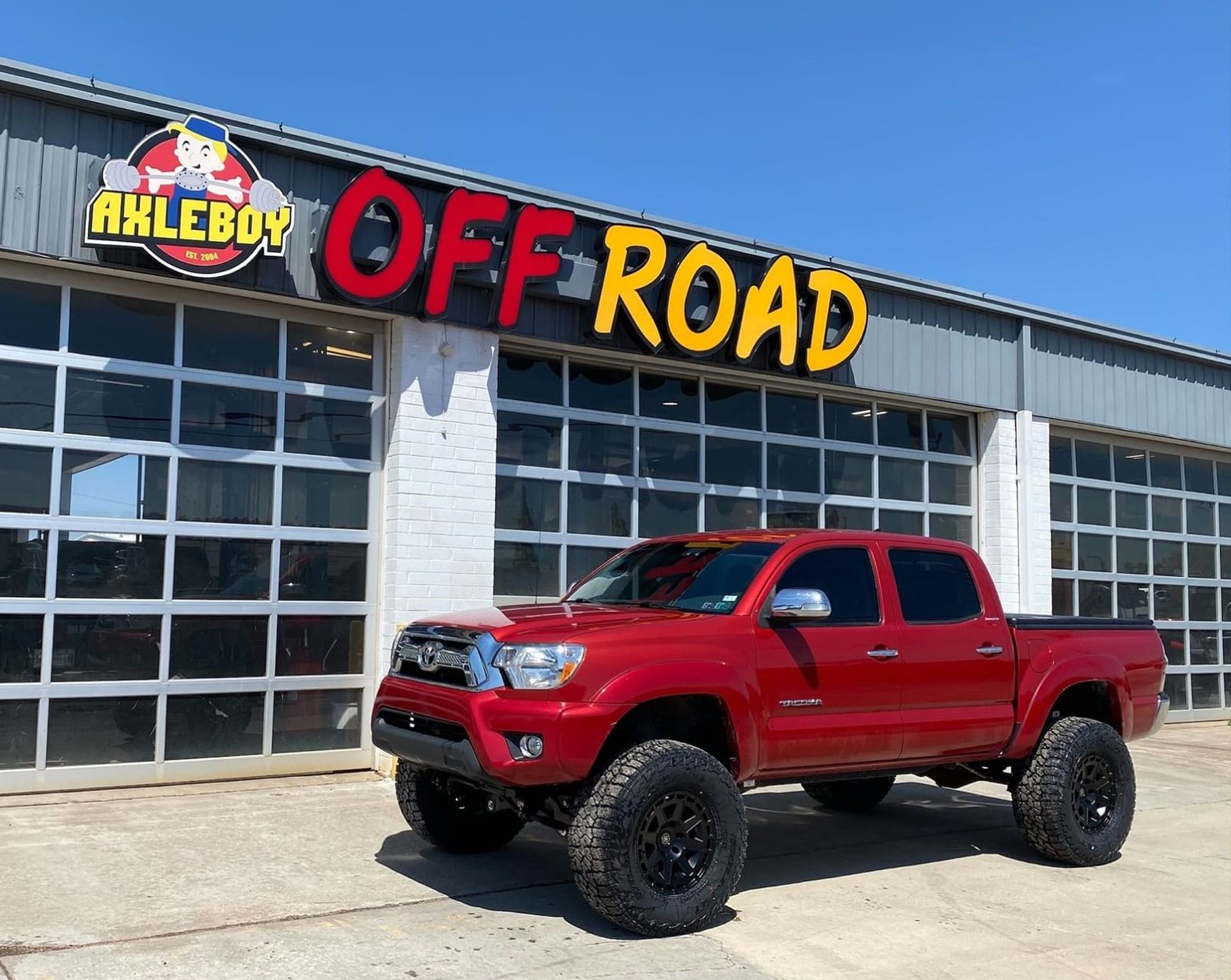A lifted red pickup truck with aggressive all-terrain tires and black wheels is parked on concrete