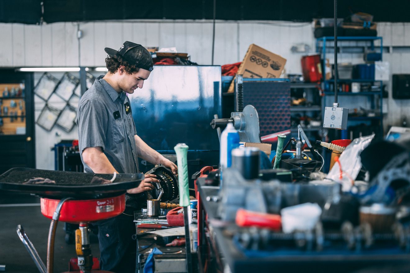 Mechanic working on a car part in a cluttered auto repair shop, wearing a cap and grey work shirt.
