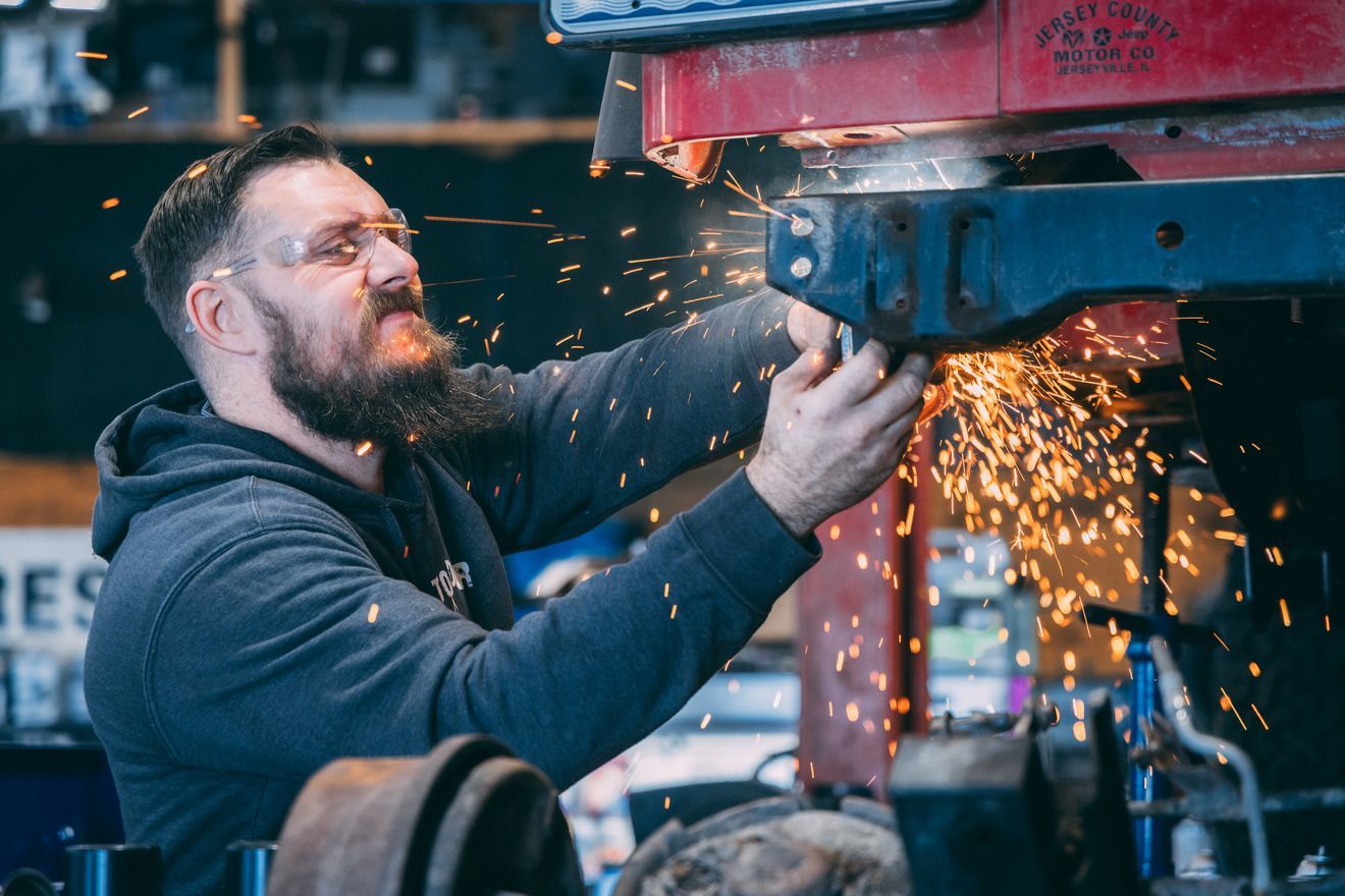 Mechanic with beard welding vehicle part, sparks flying. Wearing safety glasses, working in garage.