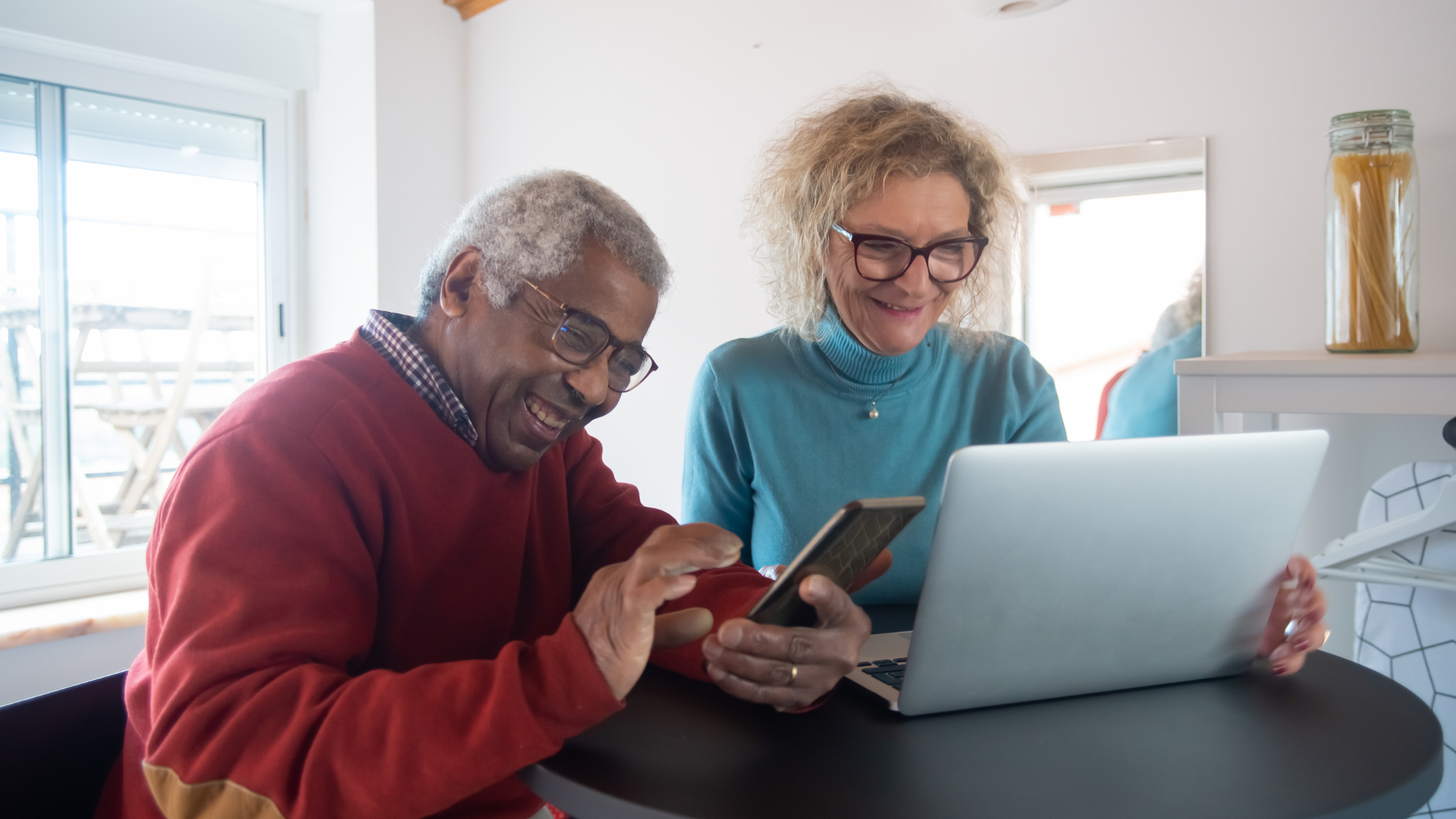 Older man on his mobile phone sitting next to an older woman on her laptop, both smiling