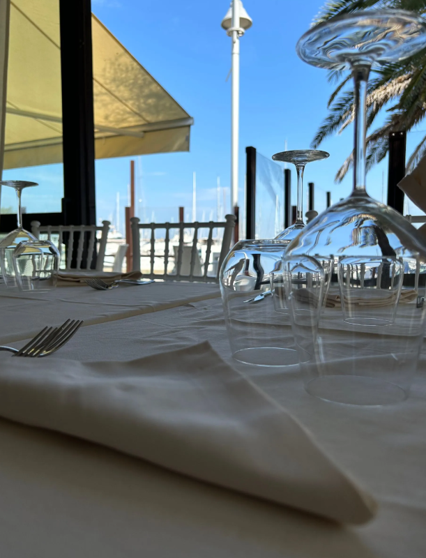 Tavolo del ristorante apparecchiato con bicchieri e tovagliolo, con vista sul porto e sul cielo azzurro.