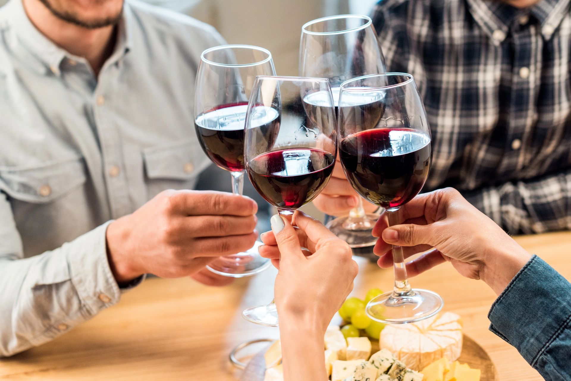 Persone che brindano insieme con calici di vino rosso, festeggiando attorno a un tavolo con formaggio e frutta.