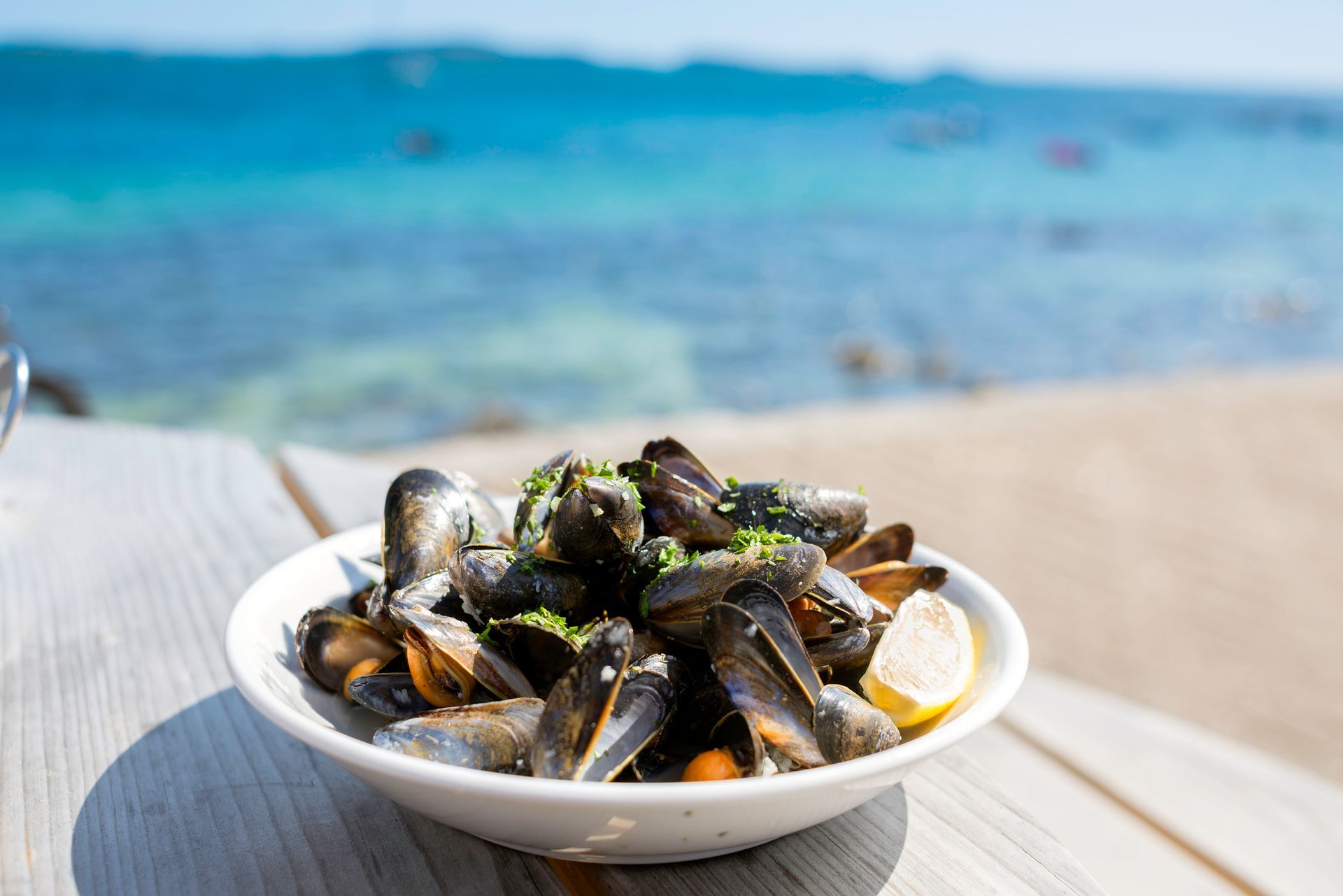 Ciotola di cozze al vapore con limone, su un tavolo in spiaggia. Oceano turchese sullo sfondo.